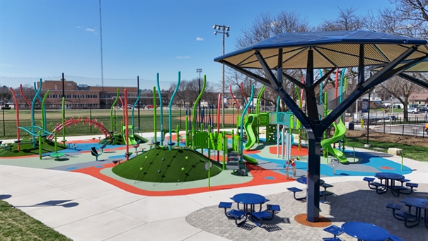 Colorful outdoor playground with climbing structures, slides, and play equipment on a soft surface, surrounded by picnic tables and a shaded pavilion.