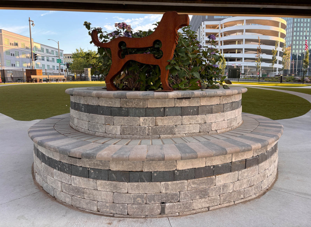 Stone planter with a cut-out metal dog shape on top, surrounded by flowers and greenery. Urban backdrop with buildings and greenery. Calm atmosphere.
