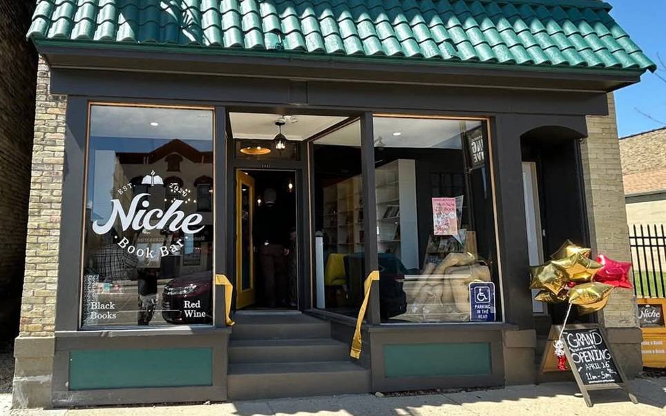 Street view of Niche Book Bar with a teal tiled roof. The entrance is decorated with a gold ribbon and balloon bouquet. A chalkboard sign announces the grand opening.