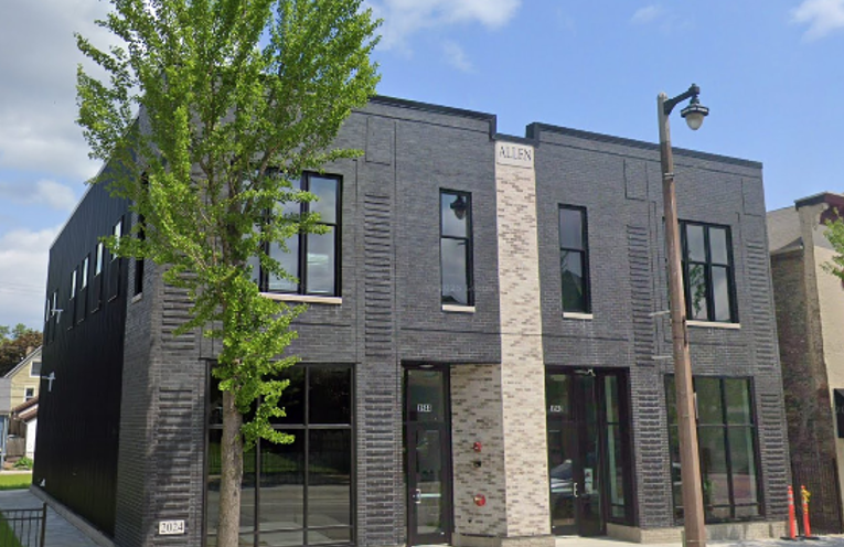 Modern two-story building with dark brick facade, large windows, and a single light-colored vertical column. A tree and streetlight are in front.