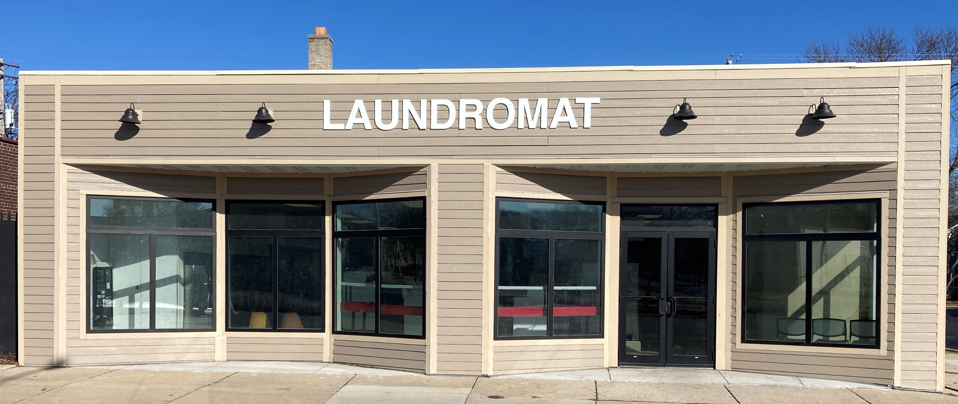 A beige laundromat building with large windows and a centered door. The word "LAUNDROMAT" is prominently displayed above. Bright daylight sets a clean, welcoming tone.