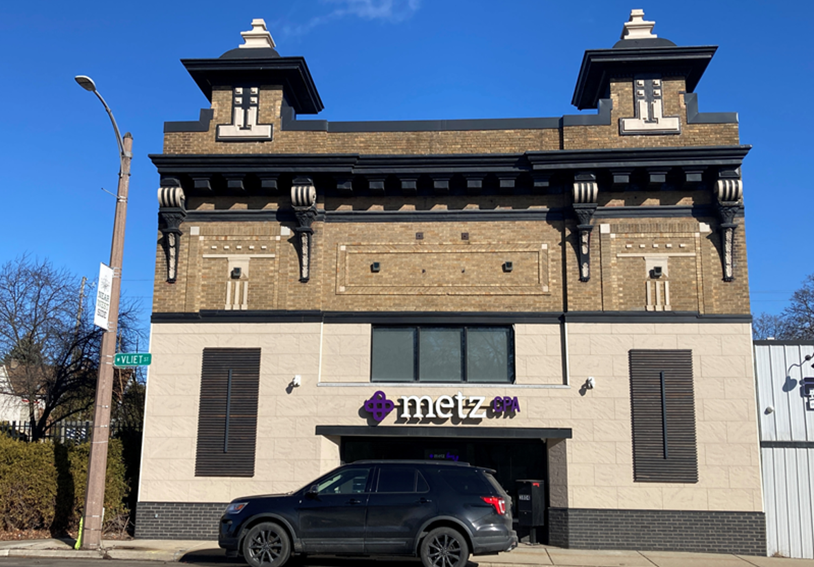 A historic-style brick building with decorative elements, housing Metz spa. A black SUV is parked in front under a clear blue sky. Calm and inviting.