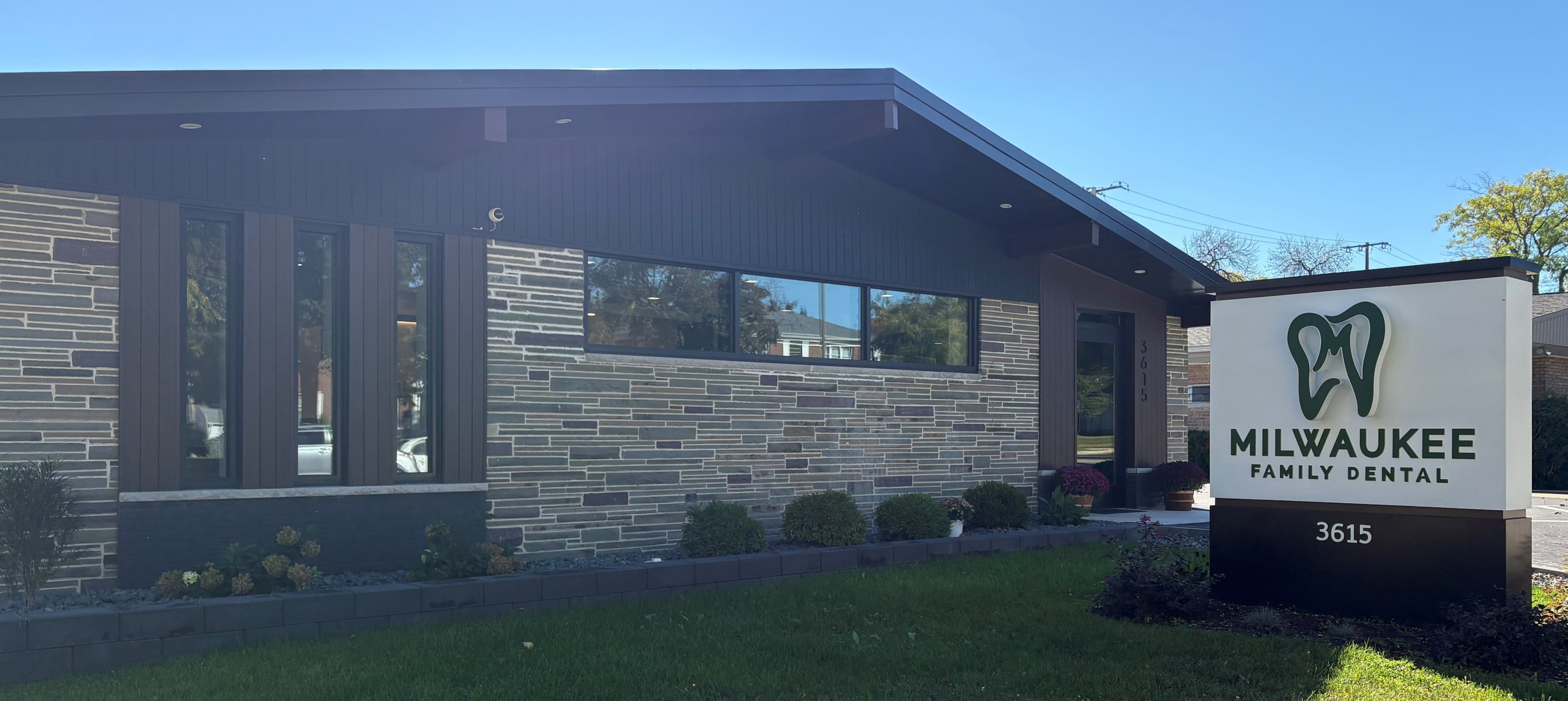 Modern dental clinic exterior with stone facade and large windows, under a clear blue sky. A sign reads "Milwaukee Family Dental" with a tooth logo.