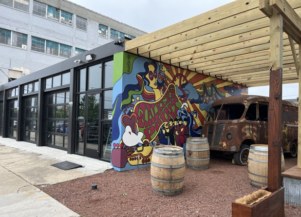 A vibrant mural covers a building wall, featuring psychedelic artwork and bold colors. Rustic barrels and a vintage van are under a wooden pergola, creating a retro atmosphere.
