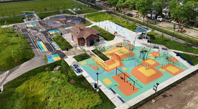 Aerial view of a park with colorful playgrounds, splash pad, basketball courts, green lawns, walking paths, and a central brick building.