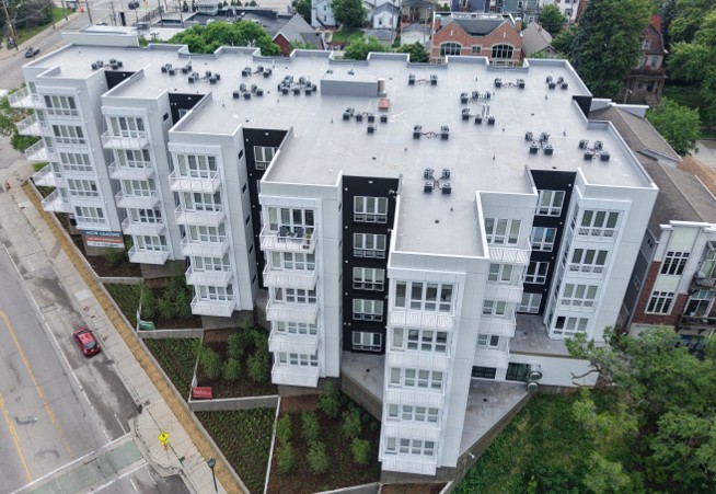 Aerial view of a modern white and gray apartment building with multiple balconies and a rooftop terrace, surrounded by trees and neighboring structures.