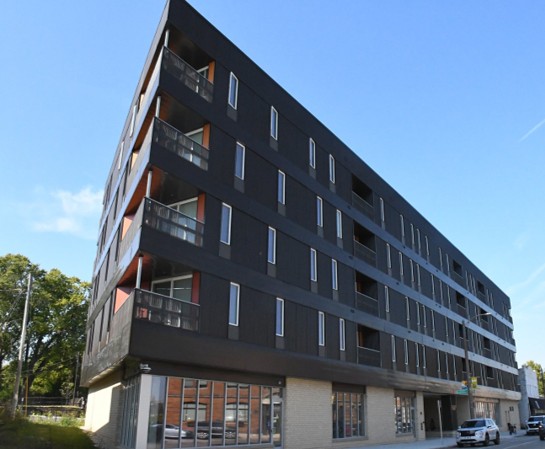 A modern, five-story apartment building with a dark facade and large windows under a clear blue sky. A car is parked at the street corner.