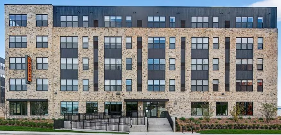 A modern, five-story brick apartment building with large rectangular windows. The entrance features steps and a ramp surrounded by greenery.