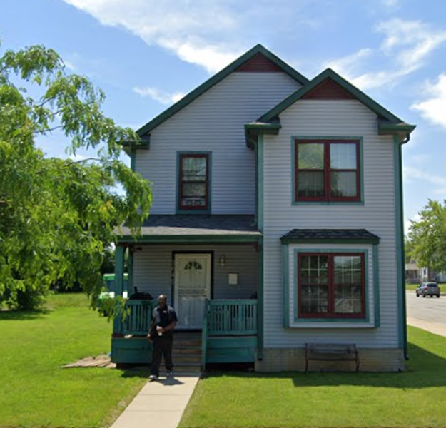 A two-story house with light gray siding, green trim, and red accents sits under a clear blue sky. A man stands in front, giving a welcoming feel.