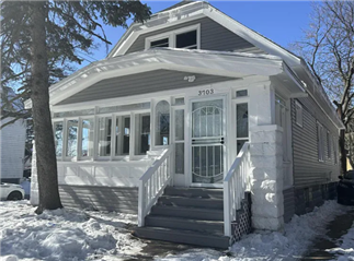 Cozy, snow-covered gray house with white trim, large front windows, and a welcoming porch. Tall trees frame the scene under a clear blue sky.