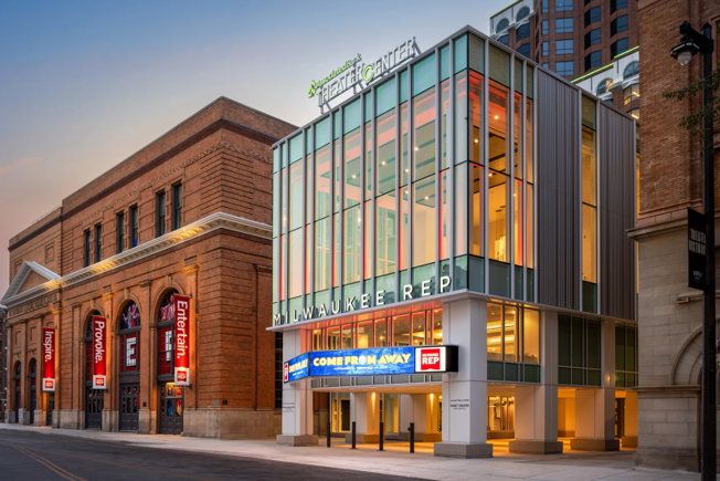 Exterior of Milwaukee Rep, a modern glass theater building with warm interior lights. Adjacent is a historic brick building. A sign advertises "Come From Away."