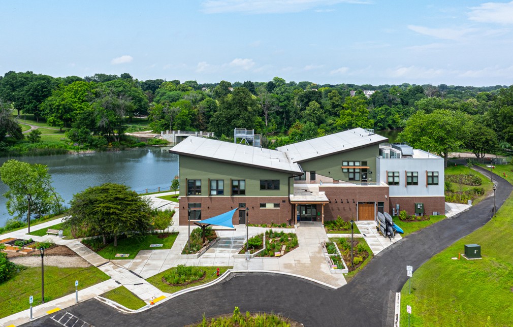 A modern building with angular roofs is situated by a tranquil river, surrounded by lush greenery. Walkways and a parking area are visible.