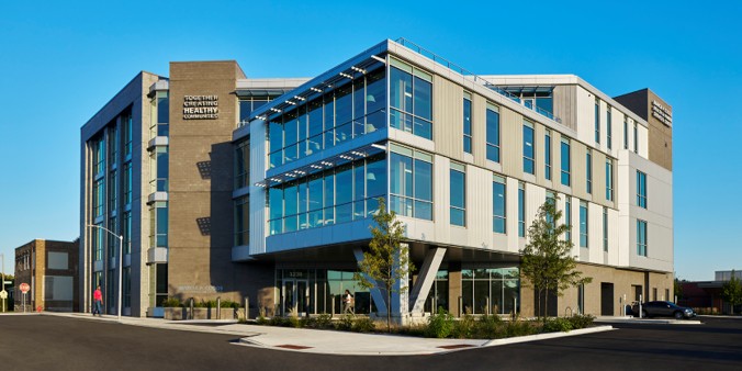 Modern three-story building with a mix of glass and concrete facade, set against a clear blue sky. Trees and people add scale and a lively atmosphere.