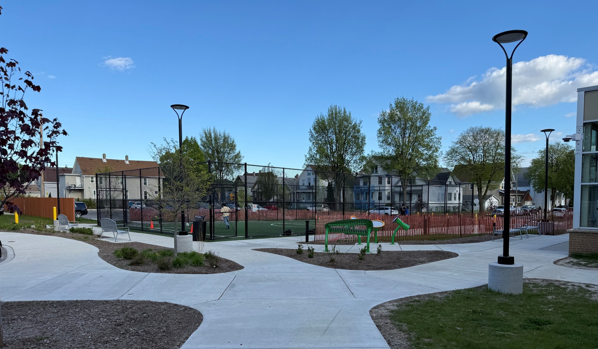A fenced sports field surrounded by trees and residential houses, under a clear blue sky. Pathways lead to the area, with lampposts and greenery.