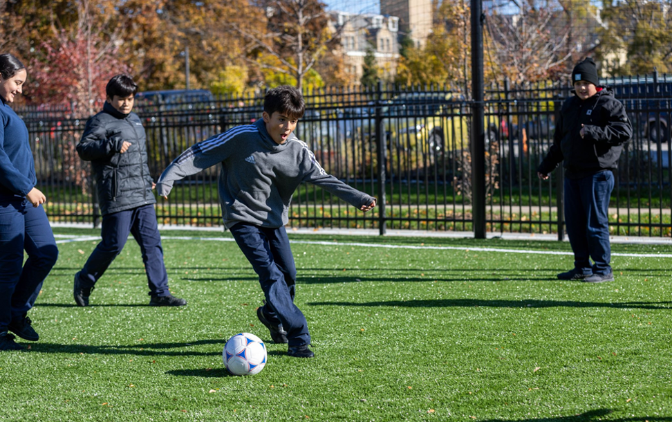 Kids playing soccer on a grassy field. One boy in a gray sweater kicks the ball, while others in jackets watch. The scene is lively and sunny.
