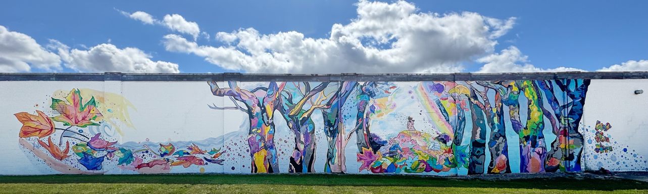 Vibrant mural on a long wall depicting colorful abstract trees and swirling leaves against a blue sky with clouds, evoking a sense of whimsy and nature.
