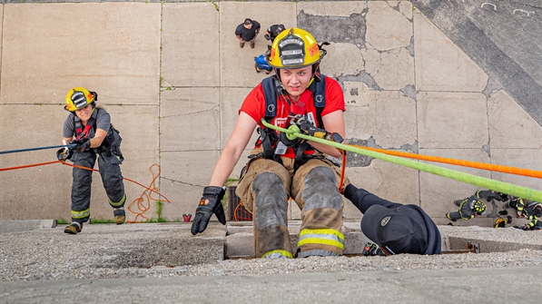 Two Junior Fire Institute students in harnesses hanging from ropes off the side of the burn tower with two onlookers on the ground in the background