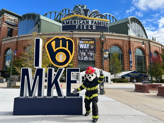 Sparky the Fire Dog in front of American Family Field next to a blue I (yellow Brewers mitt symbol) MKE sign