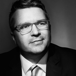 City Attorney Evan Goyke wearing glasses, a suit, and a striped tie looks slightly upward in a black and white portrait with a neutral background.