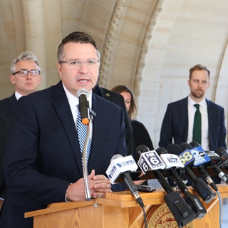 City Attorney Evan Goyke, wearing a dark suit and light blue tie, speaks at a podium with multiple microphones labeled with news logos. Several people in suits stand behind him in an arched walkway.