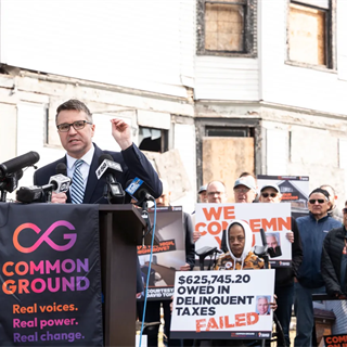 City Attorney Evan Goyke at a podium speaking to a crowd in front of a dilapidated building. Signs with messages about taxes and housing issues surround him, conveying urgency.