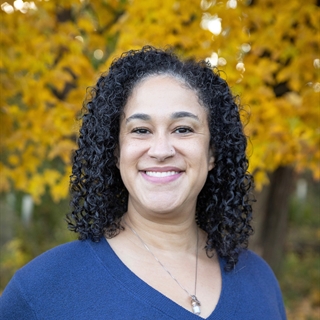 A woman smiling with curly hair in a blue top stands before vibrant yellow autumn leaves. The tone is warm and cheerful.