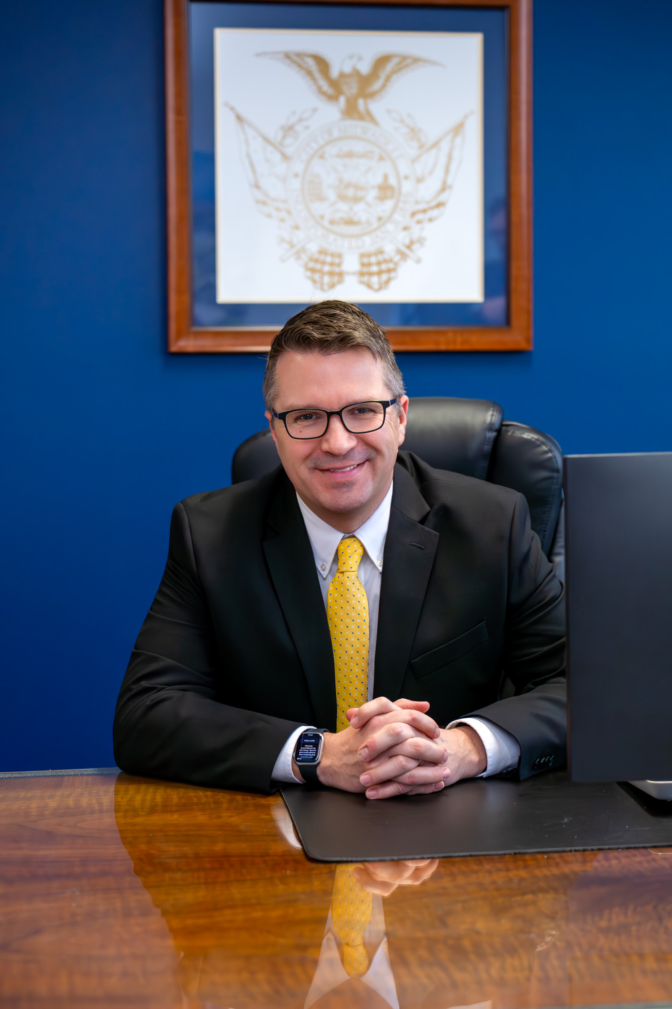 Man in a suit and yellow tie sits at a wooden desk with hands clasped, smiling confidently. Behind him, a framed emblem hangs on a blue wall.