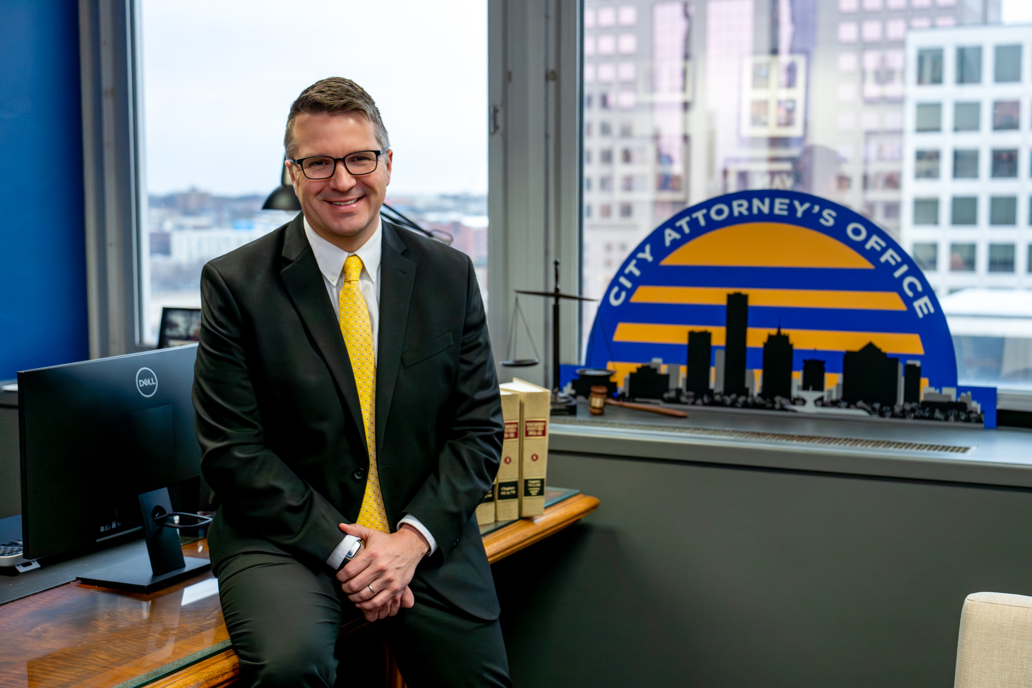 Man in a suit with a yellow tie, smiling and sitting on a desk in an office, next to books and a sign reading "City Attorney's Office" with a city skyline.