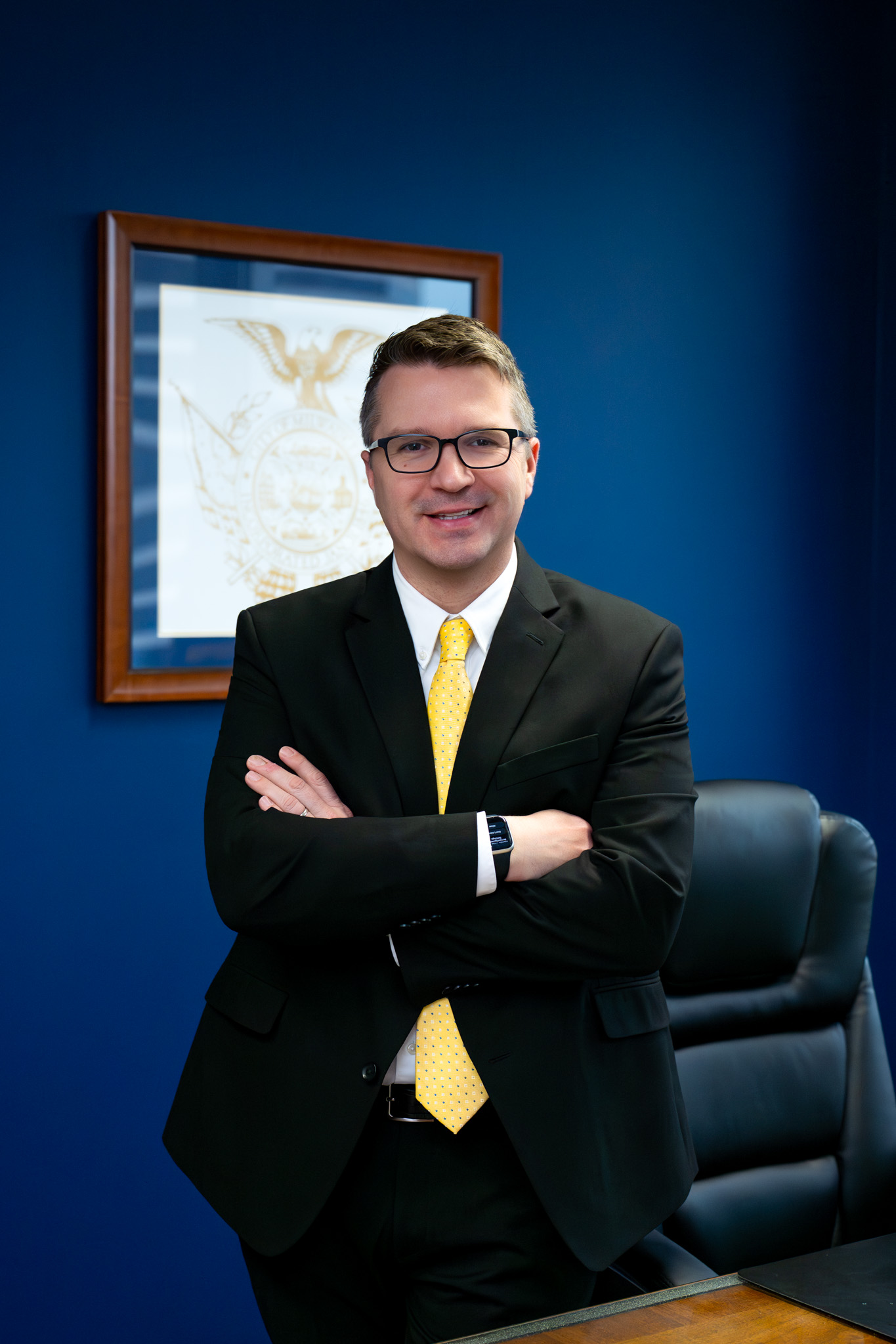 Man in a black suit with a yellow tie, standing confidently with arms crossed in an office, beside a dark blue wall with a framed certificate.