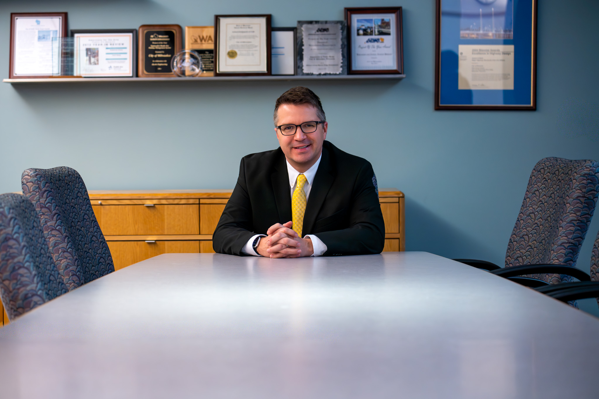 A man in a suit and yellow tie sits confidently at the head of a conference table. Behind him, a wall displays framed awards and certificates.