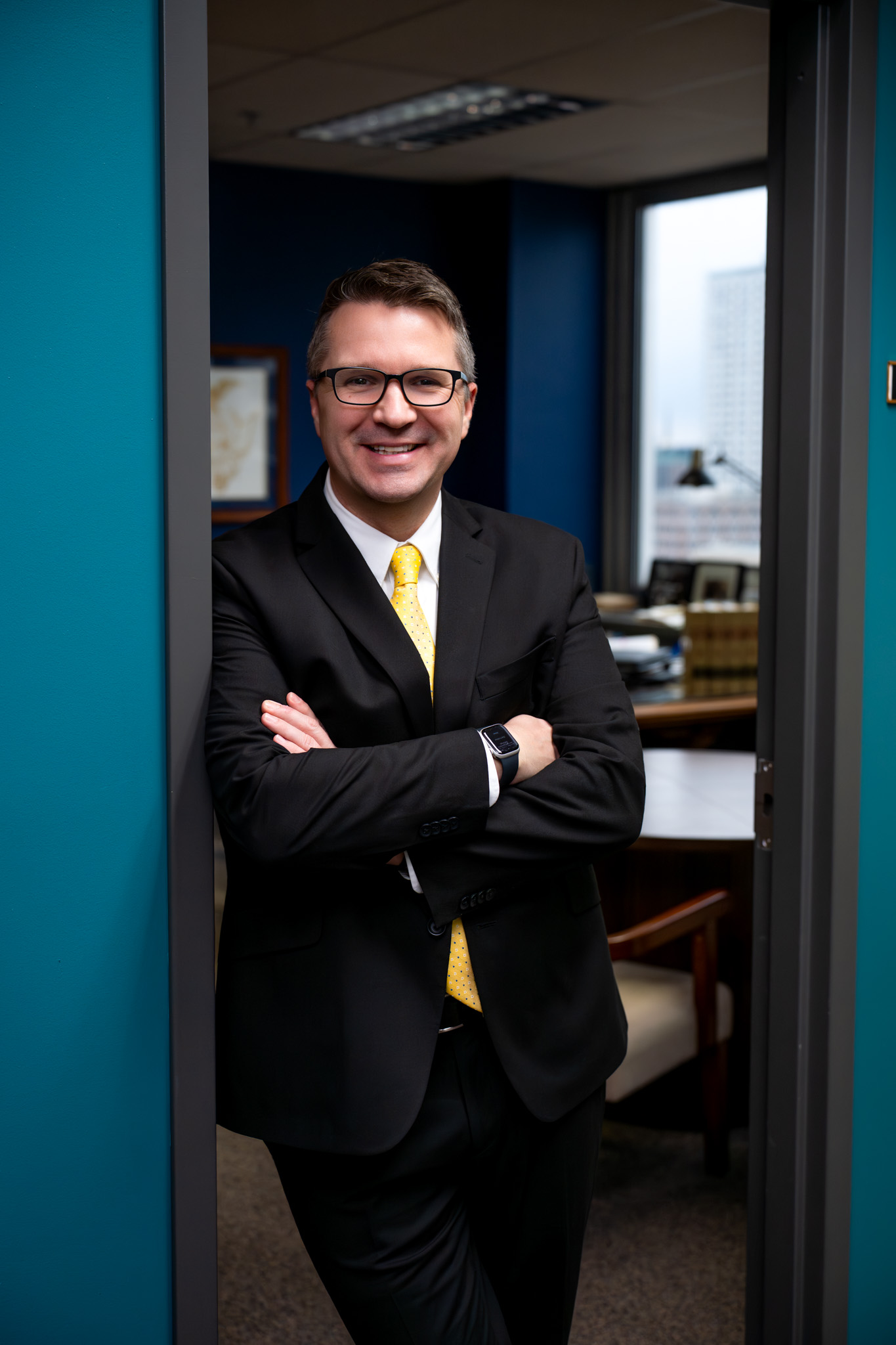 A man in a black suit and yellow tie smiles confidently, leaning against a doorframe in an office with teal walls, creating a professional atmosphere.