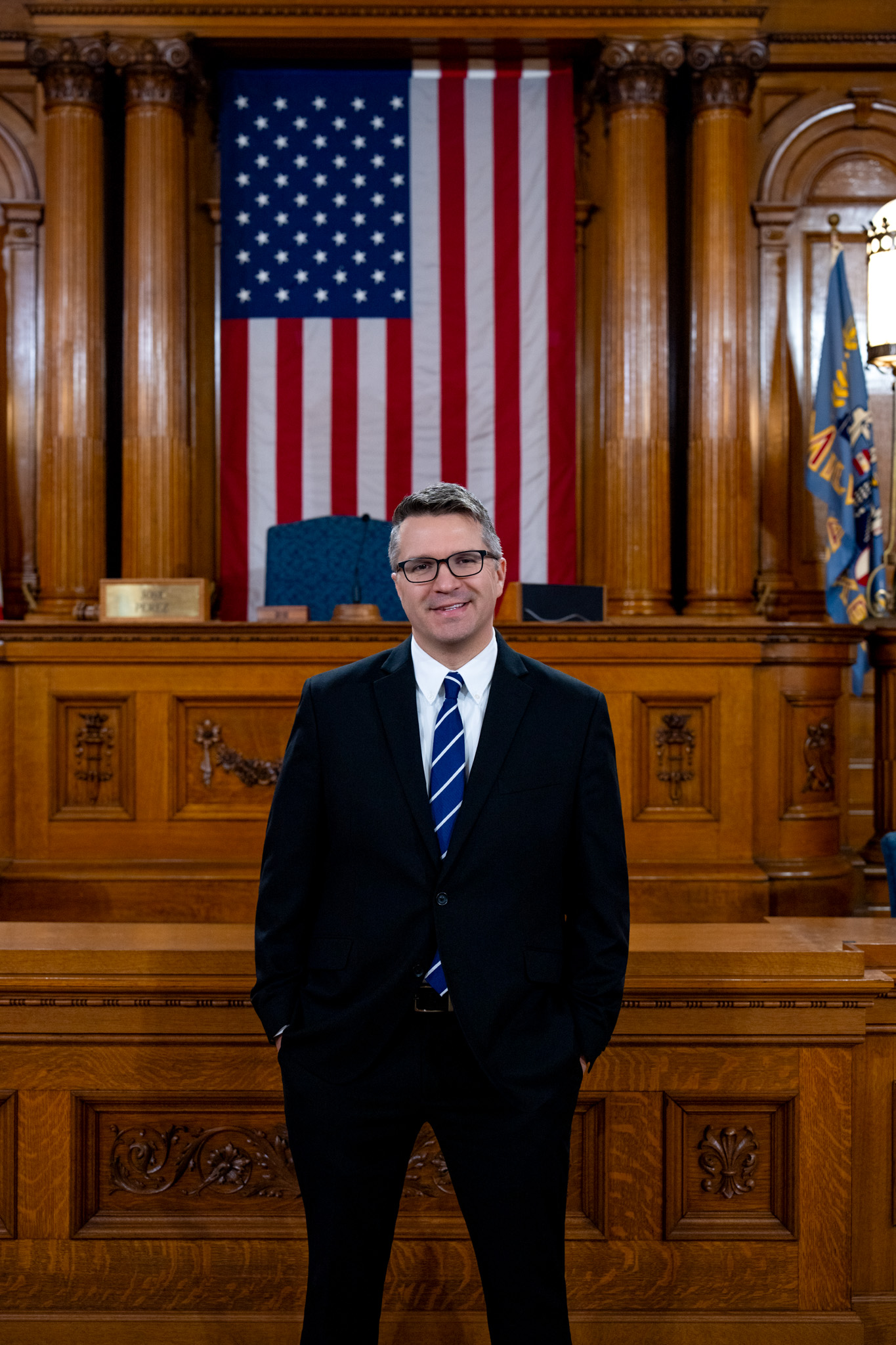 A man in a suit and striped tie stands confidently in a wood-paneled room with an American flag backdrop, evoking a formal, patriotic atmosphere.A man in a suit and striped tie stands confidently in a wood-paneled room with an American flag backdrop, evoking a formal, patriotic atmosphere.