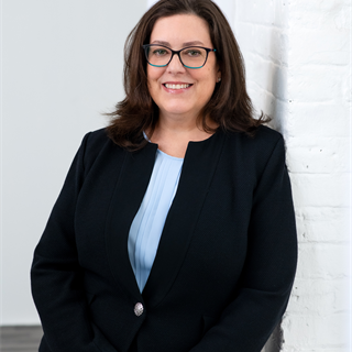 A woman with brown shoulder-length hair and glasses smiles confidently, wearing a black suit and light blue blouse standing against a white brick wall in a bright room.
