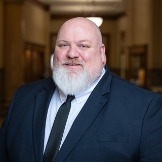 A smiling man with a white beard and bald head is wearing a navy suit and black tie. He stands in a warmly lit, elegant interior hallway.